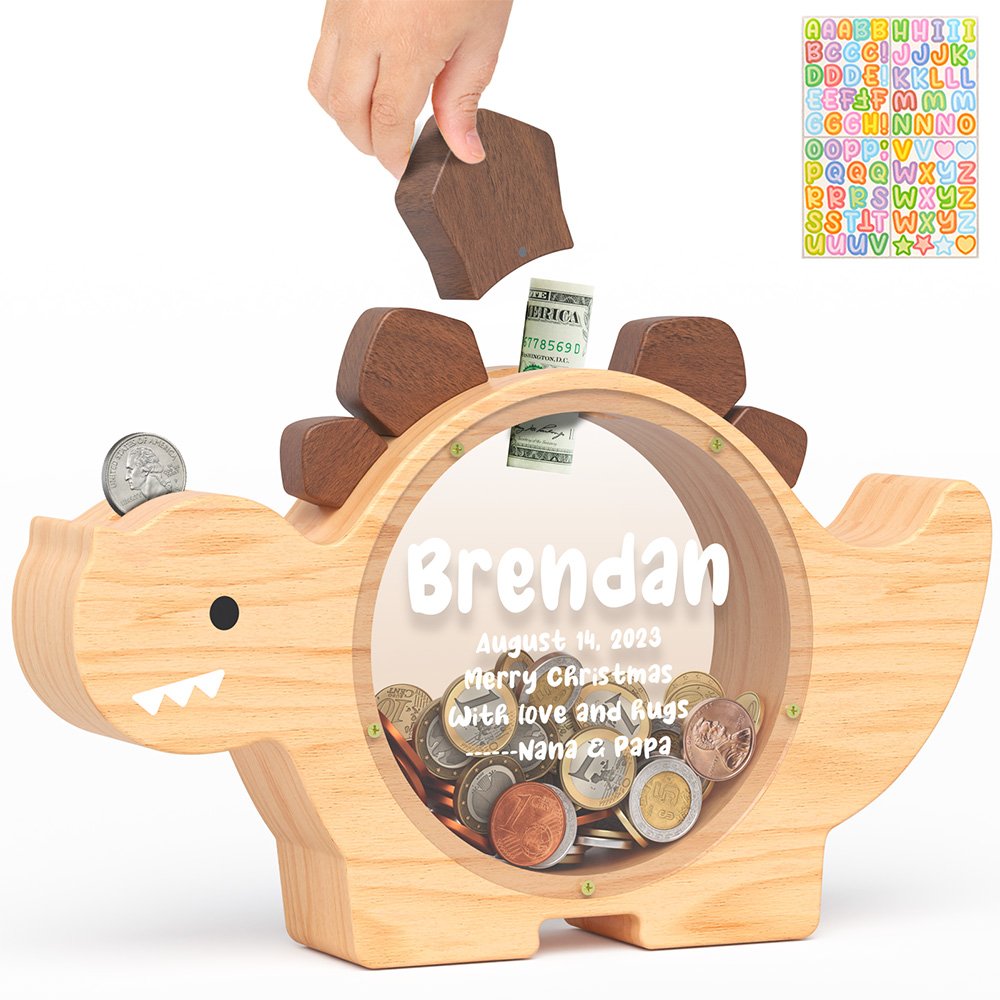 Natural wood dinosaur piggy bank with a clear acrylic front. A child's hand is dropping a coin into the top slot, complete with fun alphabet stickers for DIY decoration.
