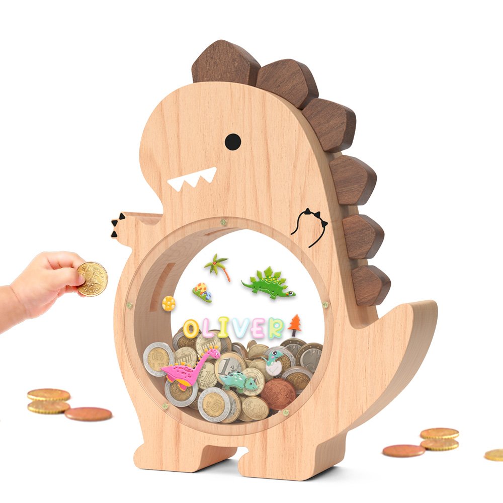 Natural wood T-Rex dinosaur piggy bank with a clear acrylic front. A toddler's hand is dropping a coin inside, decorated with colorful letter stickers spelling Oliver. A fun savings jar for boys.