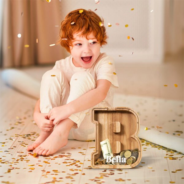 Happy child sitting with a custom wooden letter B piggy bank filled with coins. A top-rated educational toddler gift in Australia.