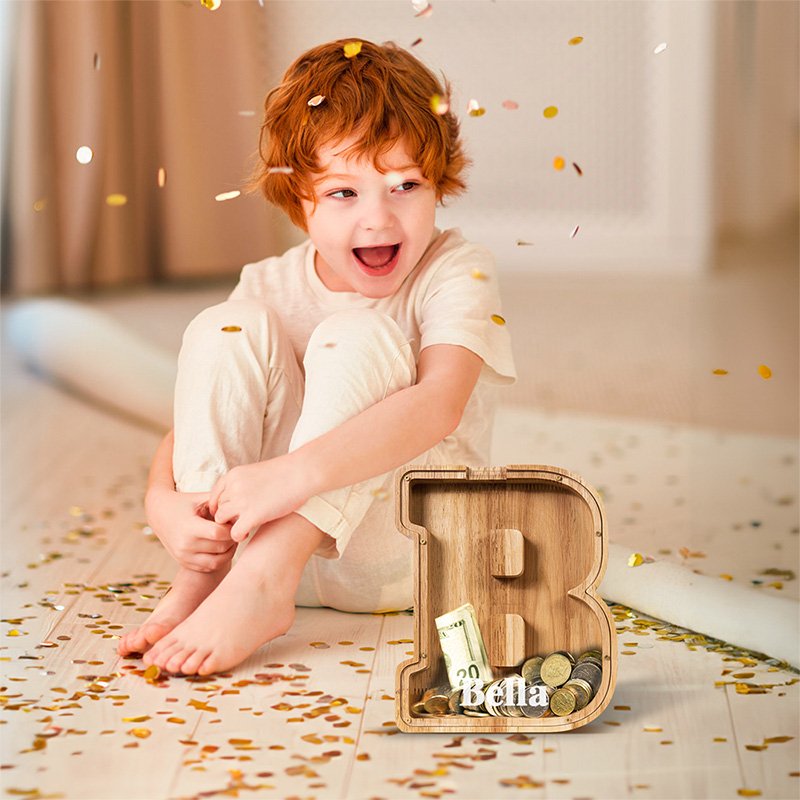 Happy child sitting with a custom wooden letter B piggy bank filled with coins. A top-rated educational toddler gift in Australia.