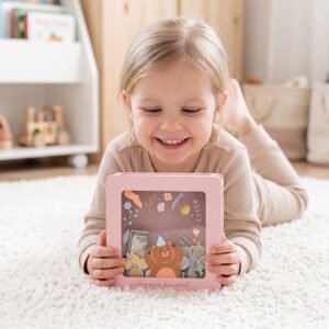 A smiling toddler girl lying on a cozy rug holding her favorite pink bear piggy bank, an ideal baby shower or birthday gift.
